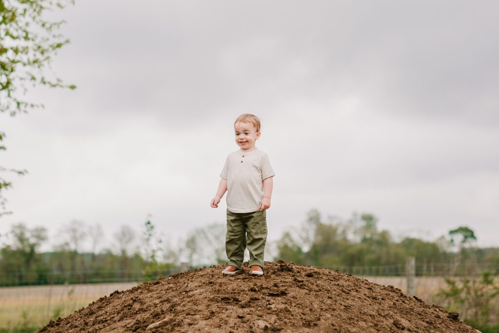 A two year old boy smiles from the top of a large dirt hill during his family photography session with Christy Rice Photography at Cooper's Knoll Farm in Pataskala, Ohio